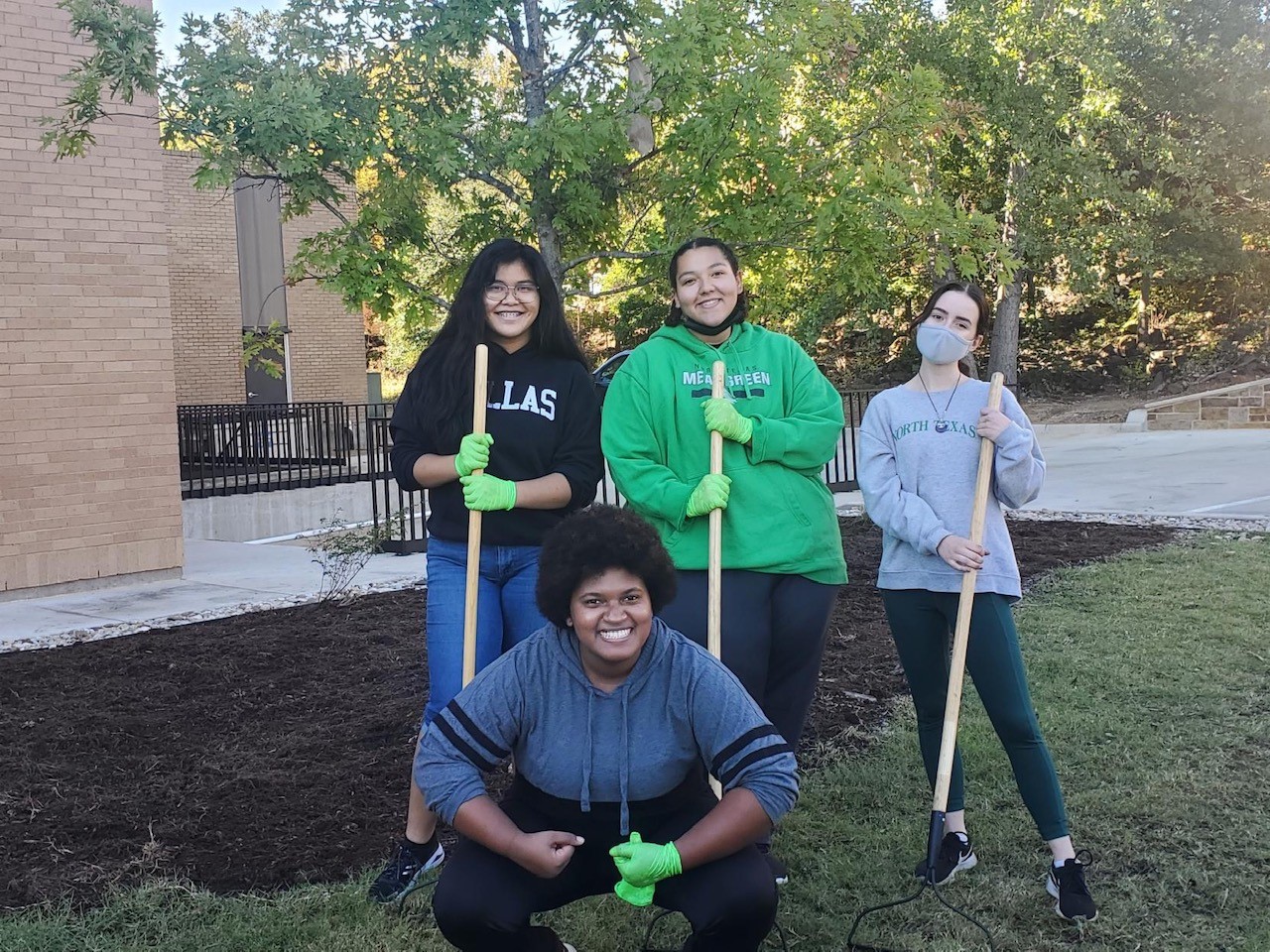 4 students stand outside with gardening rakes next to a patch of recently mulched garden with no flowers yet.