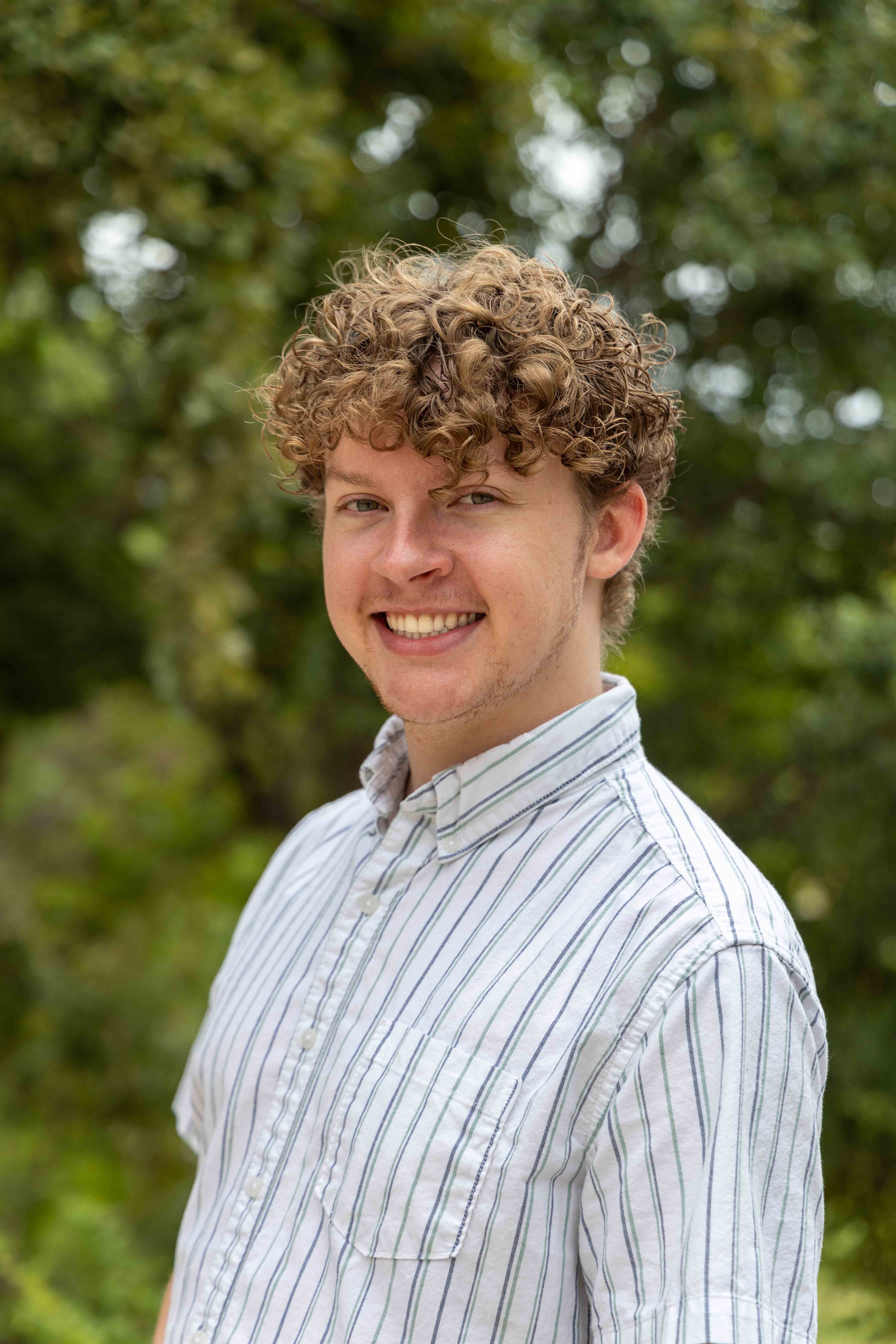 person smiling in striped button up shirt with greenery in background