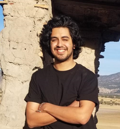 Diego standing on a mountain with a stone structure behind him wearing a black shirt and blue jeans.