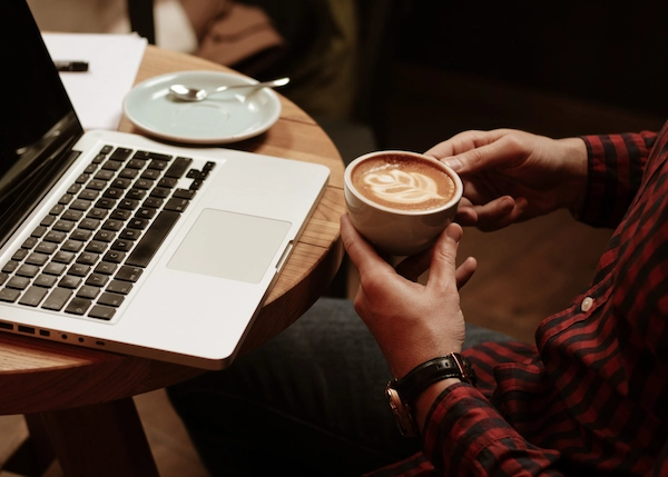 Student in a coffee shop holding a cup of coffee, sitting at a table with a laptop