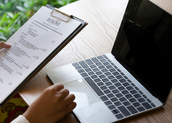 Person sitting a a desk with a laptop, holding a clipboard with a resume.
