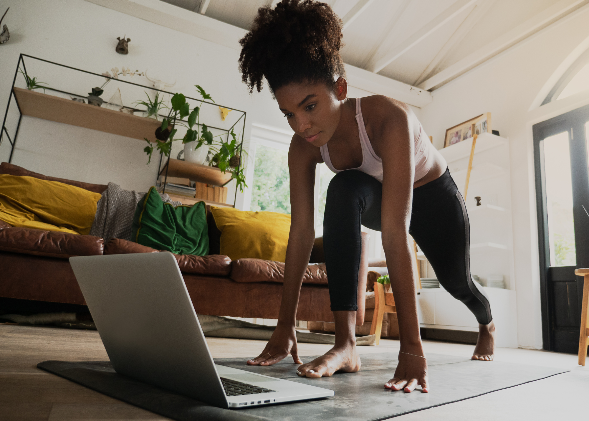 Photo of a woman stretching on a yoga mat with a laptop on the ground in front of her.
