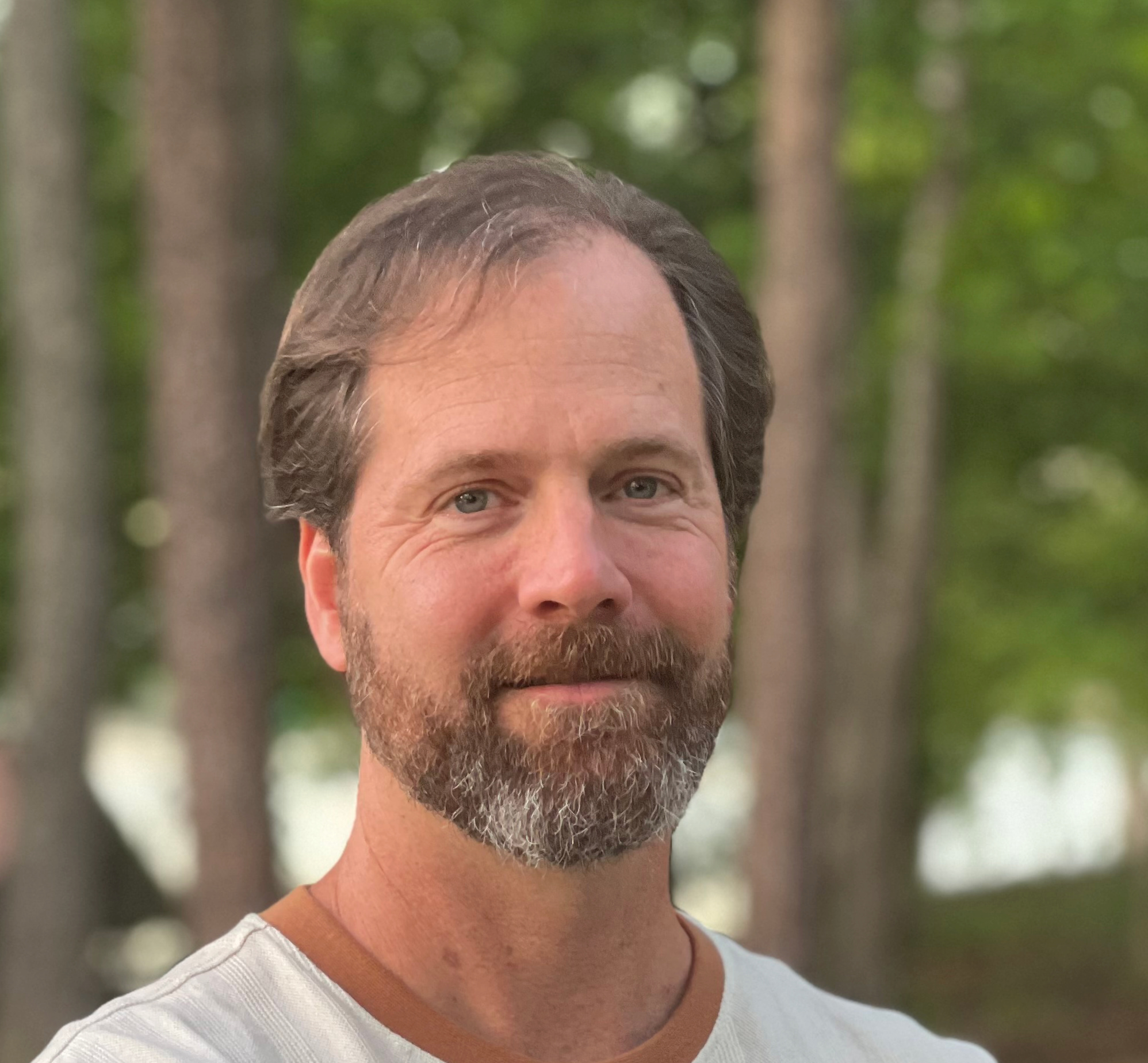 A person with short, light brown and grey hair and neatly trimmed facial hair standing outdoors in a wooded area with tall trees and green foliage.