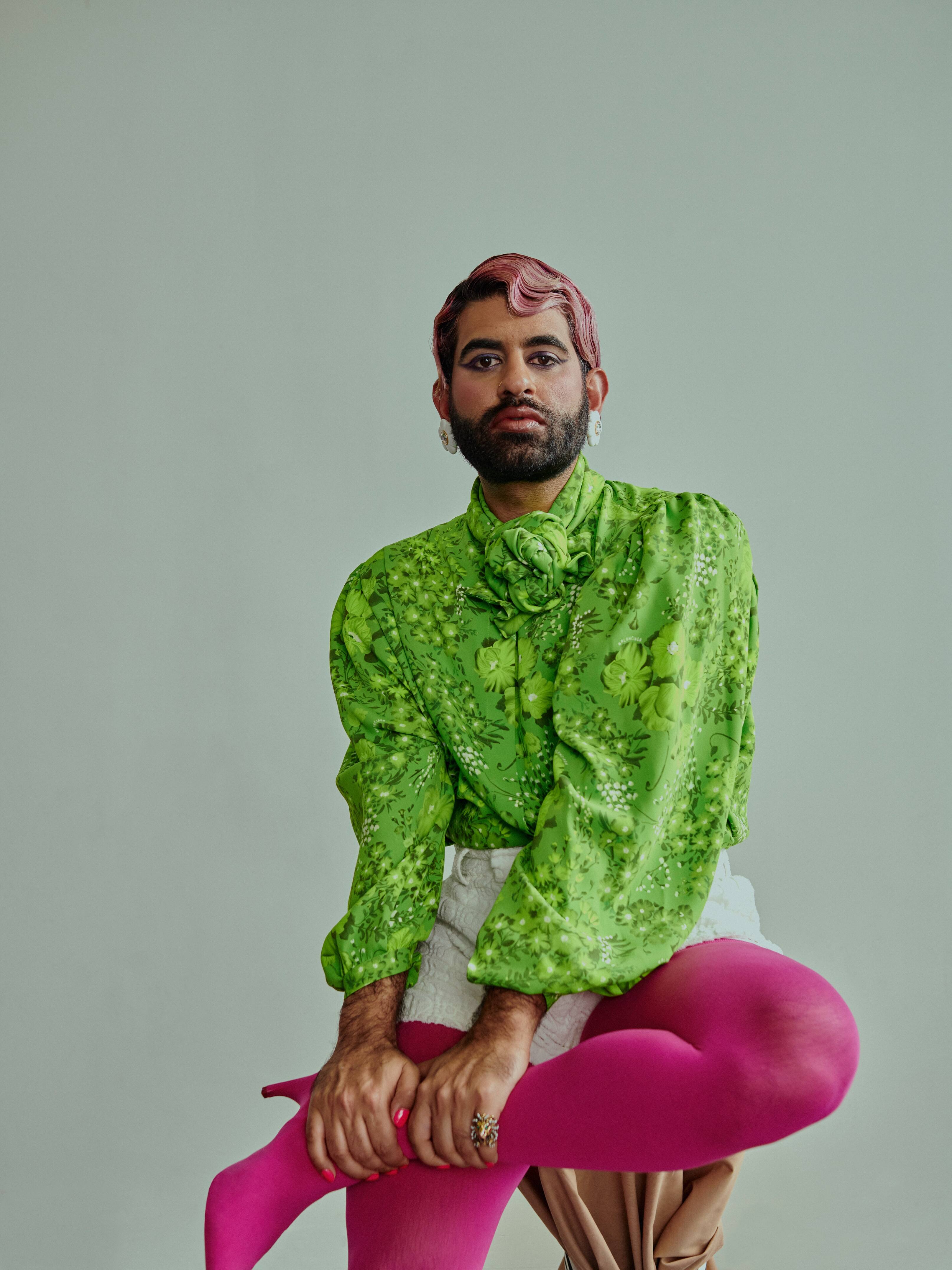 ALOK ALOK stands against a plain, light-colored background. Their face is blurred. They are wearing a bright green, long-sleeved blouse with floral patterns, paired with white shorts and vibrant pink tights.