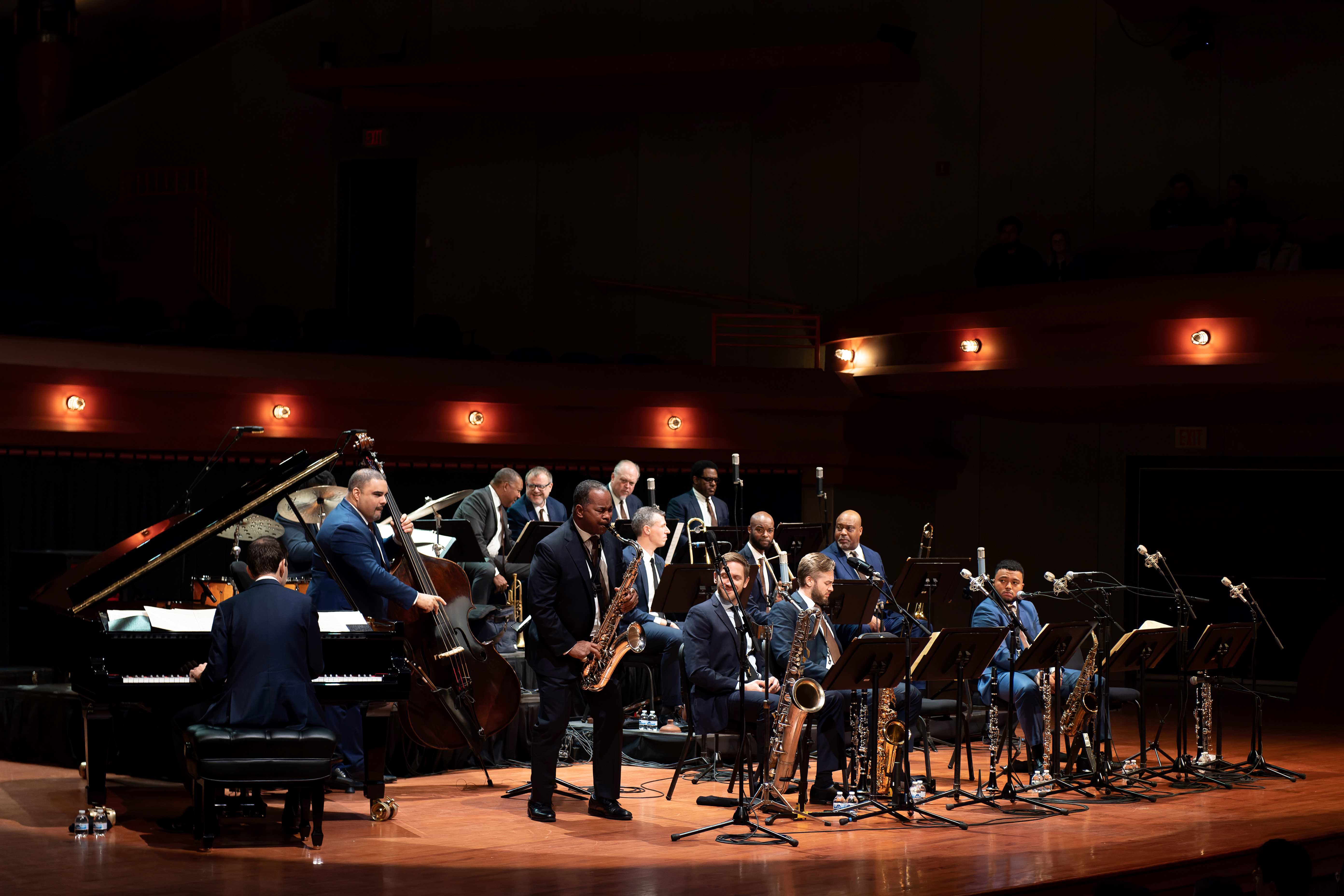 Seraph Brass with the UNT Wind Orchestra A jazz band performs on a well-lit stage in an auditorium. The ensemble includes musicians playing a grand piano, double bass, saxophones, trumpets, and trombones. They are arranged in a semi-circle, each with a music stand in front of them. All performers are dressed in formal attire. The stage lighting is warm, and the background shows the auditorium’s architectural details and wall-mounted lighting fixtures.