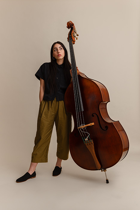 full body image of musician Mali Obomsawin with long dark hair standing with an upright bass