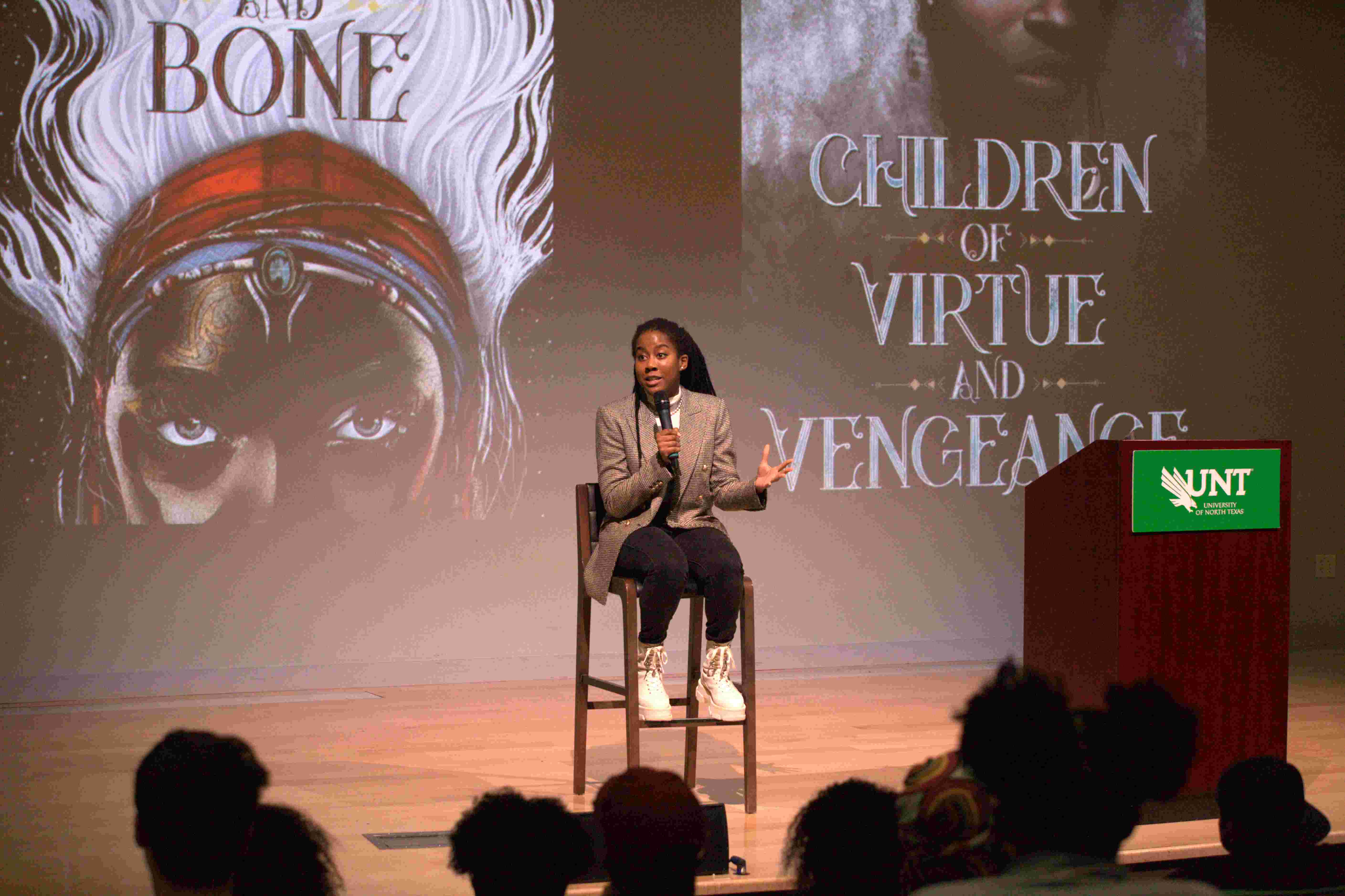 Tomi Adeyemi Tomi Adeyemi sitting on stage speaking into a microphone with her books displayed on a screen behind her.